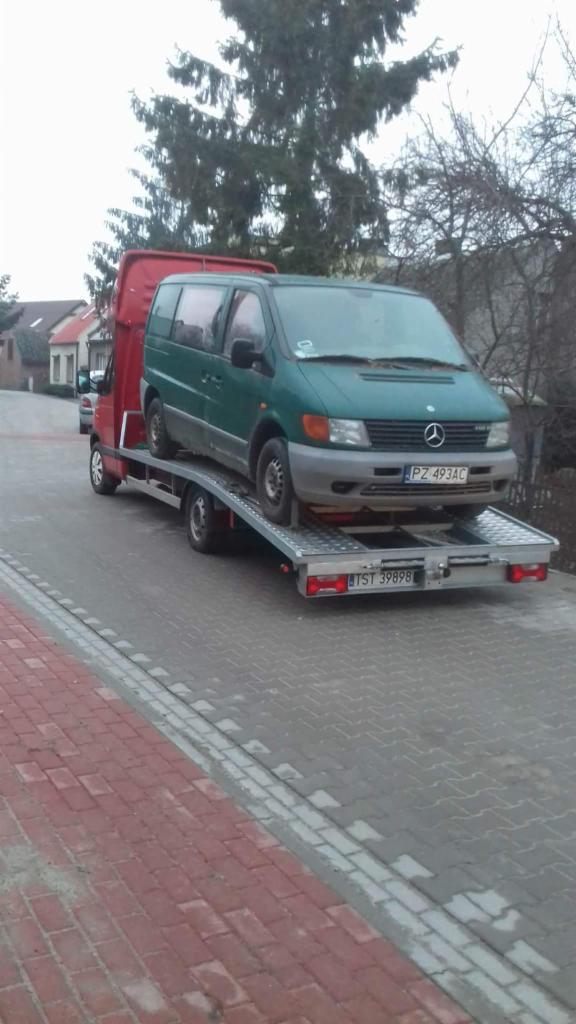 Zielony Mercedes Vito na srebrnej lawecie z czerwoną kabiną, zaparkowany na chodniku z kostki, z widocznym numerem rejestracyjnym lawety i częściowo widocznym numerem rejestracyjnym samochodu,...