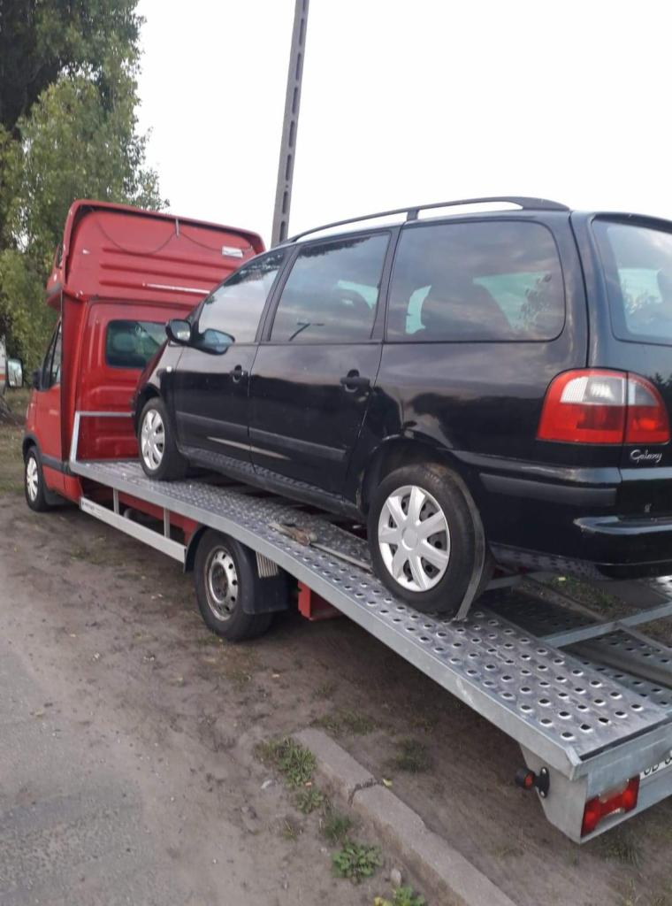 Czerwona laweta z czarnym samochodem osobowym marki Ford Galaxy na metalowej platformie, widziana z boku na tle drzew i słupa energetycznego.