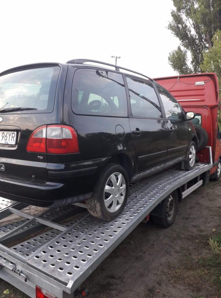 Czarny samochód Ford Galaxy kombi z bagażnikiem dachowym, z widoczną tablicą rejestracyjną 96YR, znajduje się na metalowej platformie lawety koloru srebrnego, która jest przymocowana do czerwonego...