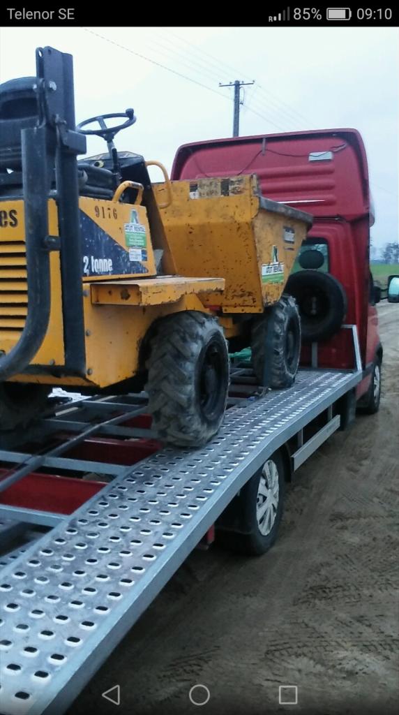 Żółta wywrotka budowlana 2-tonowa umieszczona na platformie lawety z najazdami; widoczny fragment czerwonej kabiny samochodu ciężarowego.