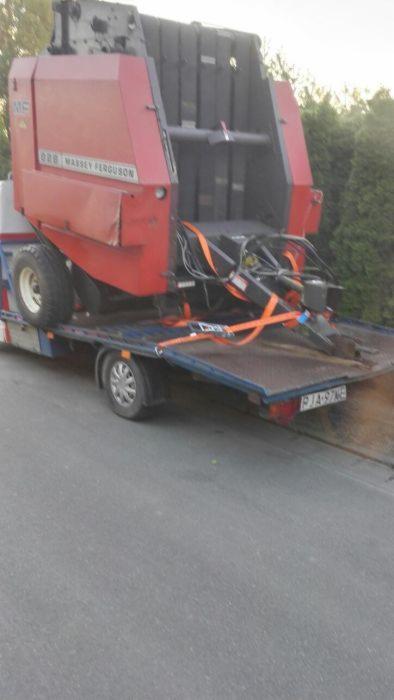 Czerwona prasa rolnicza Massey Ferguson zabezpieczona pomarańczowymi pasami na niebieskiej lawecie z widoczną tablicą rejestracyjną, drzewa w tle.