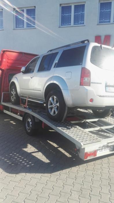 Srebrny SUV marki Nissan na lawecie w Obornikach, z widocznym czerwonym przodem pojazdu transportowego i budynkiem w tle.