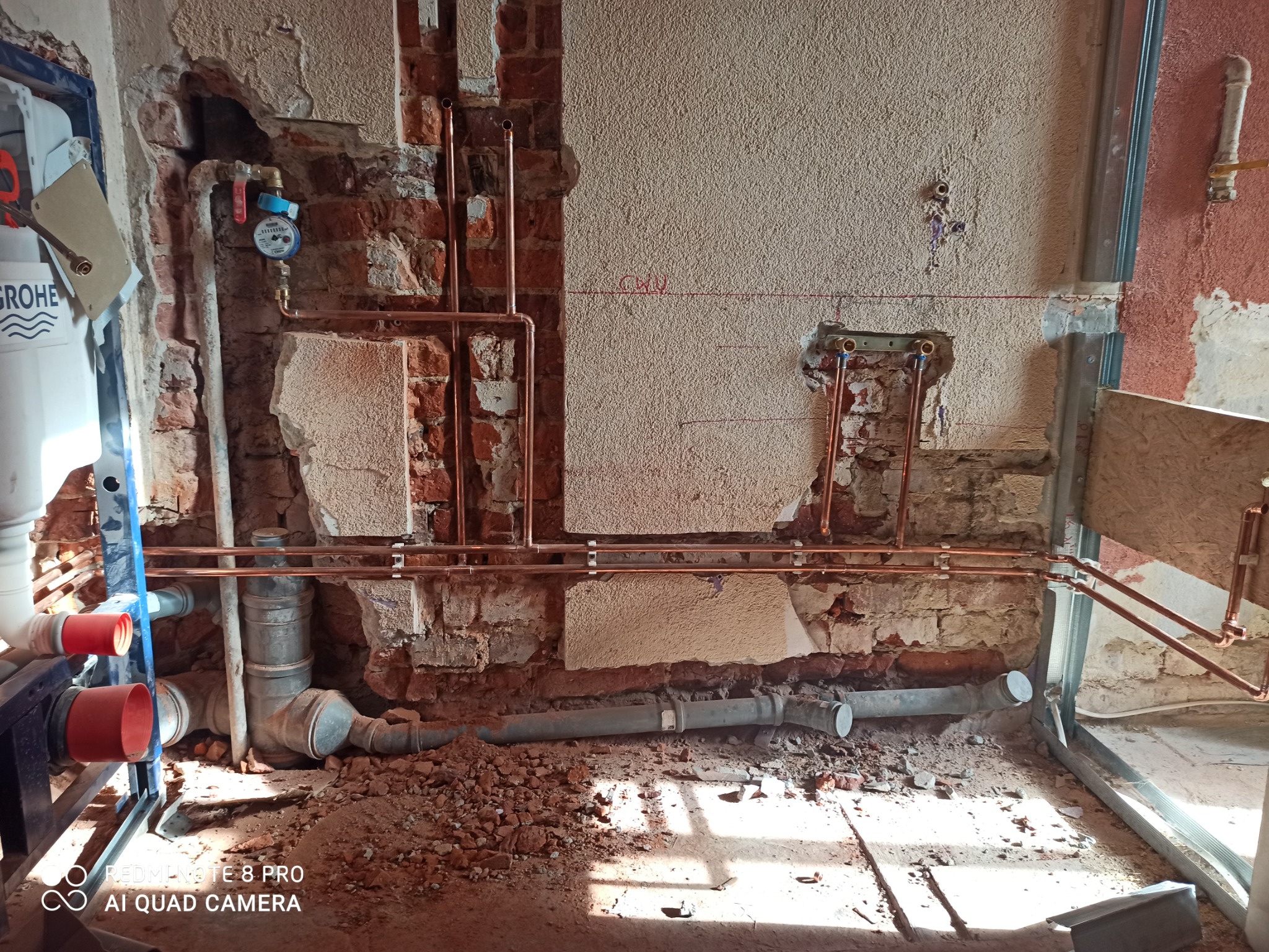 Surowe wnętrze łazienki w trakcie remontu: odsłonięte rury miedziane instalacji wodnej na tle ceglanej ściany z fragmentami tynku, widoczny wodomierz i elementy mocujące stelaż podtynkowy.