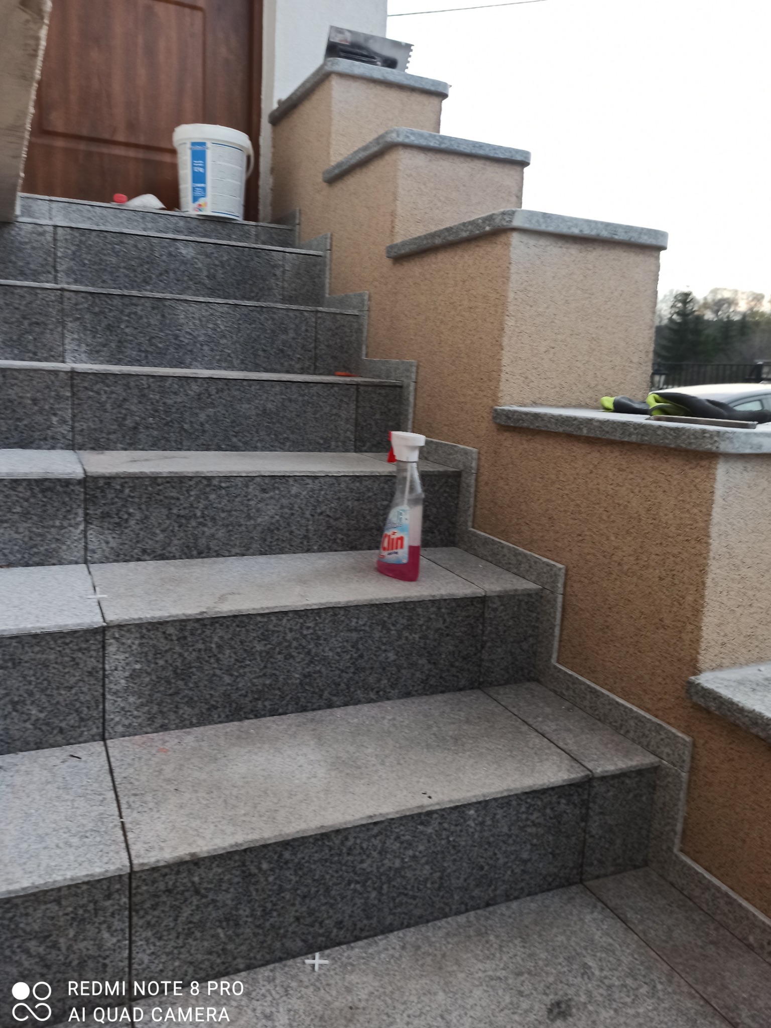 Schody zewnętrzne wyłożone szarą, granitową płytką, widoczny biały pojemnik z farbą, butelka płynu czyszczącego i rękawice robocze.