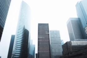 Widok z dołu na nowoczesne biurowce w centrum miasta, dominują szklane fasady i stalowe konstrukcje, niebo jest jasne, a perspektywa podkreśla wysokość budynków.