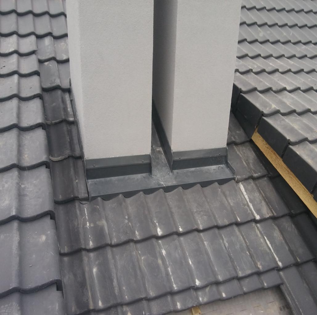 Szczegółowe ujęcie szarej dachówki ceramicznej wokół podstawy komina z obróbką blacharską w kolorze grafitowym.
