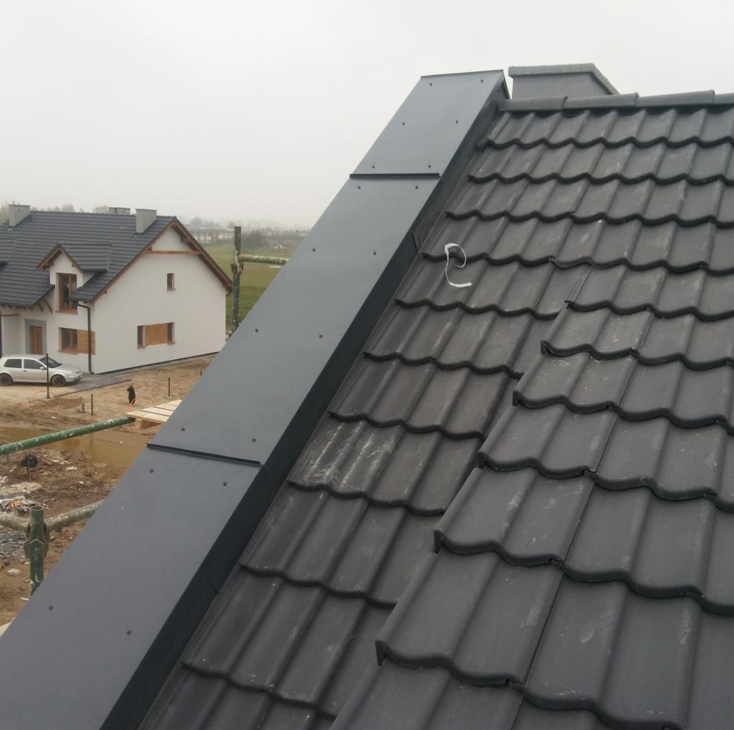 Fragment dachu z czarną dachówką ceramiczną i obróbką blacharską w kolorze grafitowym, widok z góry na tle rozmytego krajobrazu i domu w budowie.
