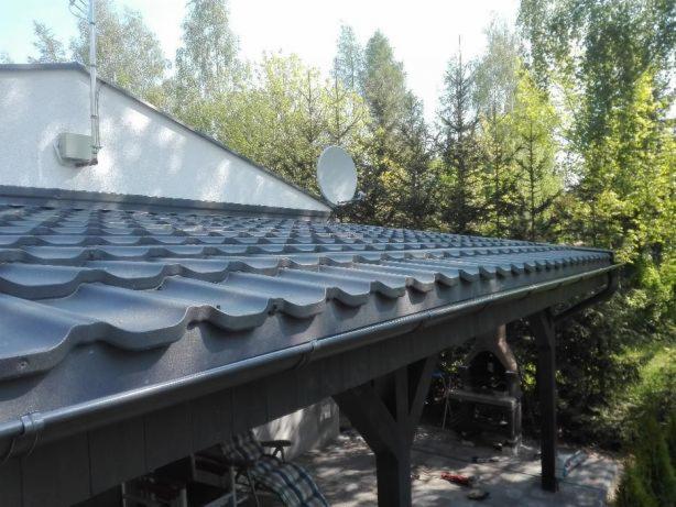 Szary dach z blachodachówki z zamontowaną anteną satelitarną na tle zielonych drzew.