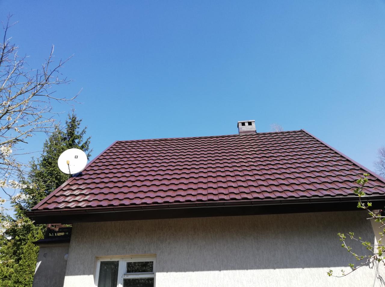 Ciemnobrązowy dach z blachodachówki trapezowej, widoczny komin z cegły, antena satelitarna, jasna elewacja budynku i błękitne niebo.