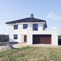 Nowoczesny, dwukondygnacyjny dom z białymi ścianami, ciemną stolarką okienną i garażem w bryle budynku, widoczny w słoneczny dzień z niewielkim kopcem piasku na trawniku.