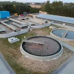 Widok z lotu ptaka na kompleks oczyszczalni ścieków z okrągłymi zbiornikami i prostokątnymi reaktorami, otoczony zielenią i budynkami technicznymi, z niebieskim kontenerem na pierwszym planie.