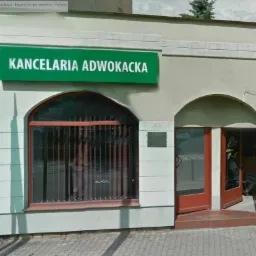 Fasada budynku z zielonym szyldem 'Kancelaria Adwokacka' nad oknem z kratami i otwartymi drzwiami wejściowymi.