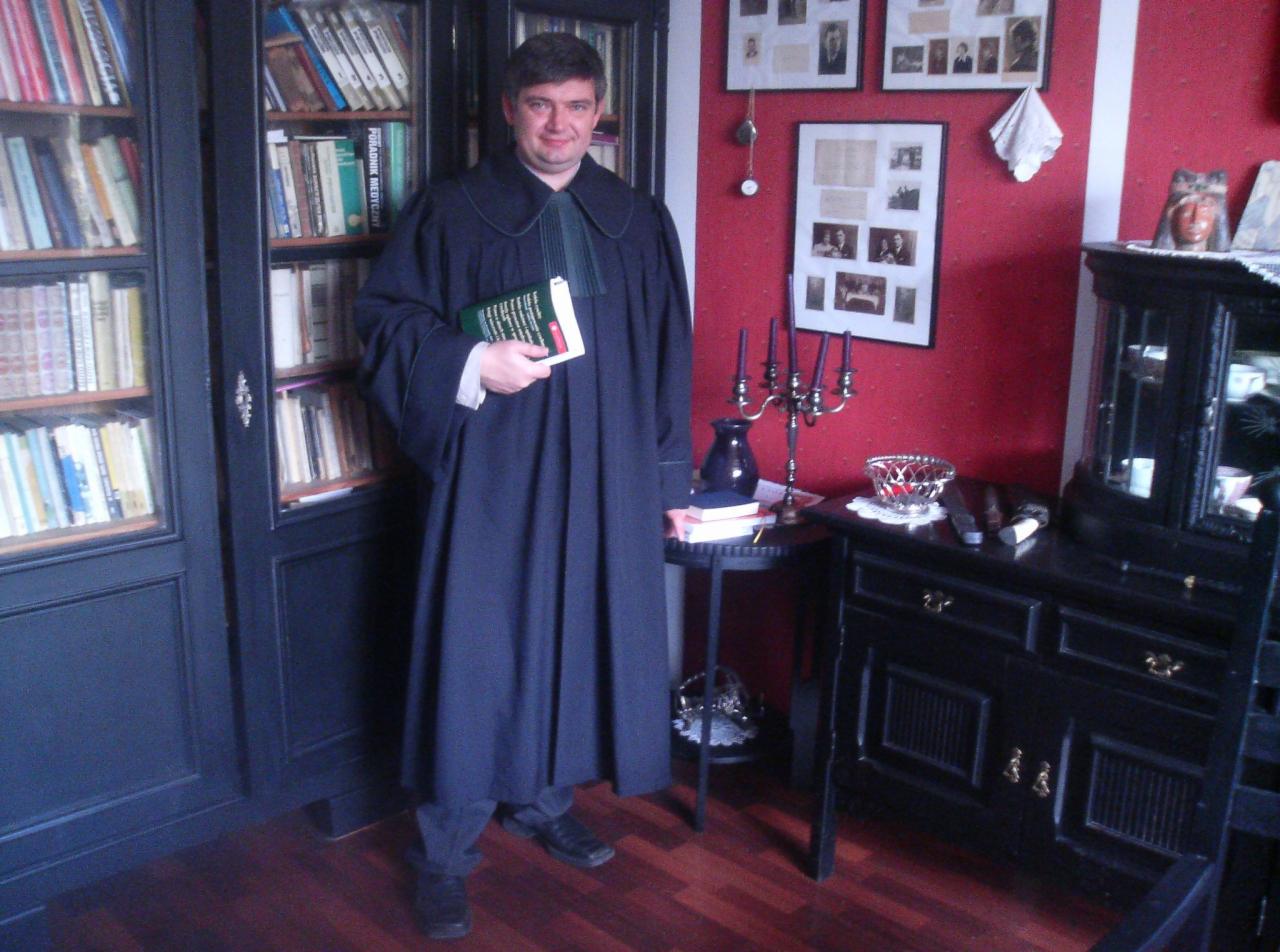 Portret mężczyzny w todze adwokackiej, trzymającego w ręku książkę przed regałem z książkami i antykami.