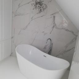 HYDROMASTERS - Nowoczesna łazienka na poddaszu z wolnostojącą białą wanną, chromowaną baterią podłogową i marmurowymi płytkami na ścianie. W tle widoczna szafka w zabudowie i ozdobny żyrandol.