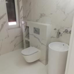 HYDROMASTERS - Nowoczesna łazienka wykończona białymi płytkami imitującymi marmur, wisząca toaleta z ukrytą spłuczką, umywalka cylindryczna na postumencie i okno z brązowymi roletami.