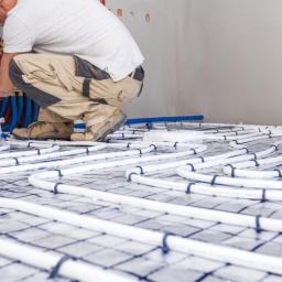 HYDROMASTERS - Instalator w kasku łączy rury ogrzewania podłogowego do rozdzielacza w surowym stanie; widoczne białe rury na siatce, czerwone i niebieskie rury doprowadzające wodę, oraz robocze spodnie z brudnymi...