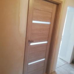 HYDROMASTERS - Zamontowane, drewniane drzwi wewnętrzne z trzema poziomymi, mlecznymi szybkami w ościeżnicy; widoczna surowa ściana w tle i podłoga imitująca drewno.