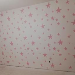 HYDROMASTERS - Ściana pomalowana w biało-różowe gwiazdki w pokoju, widoczne gniazdka elektryczne.