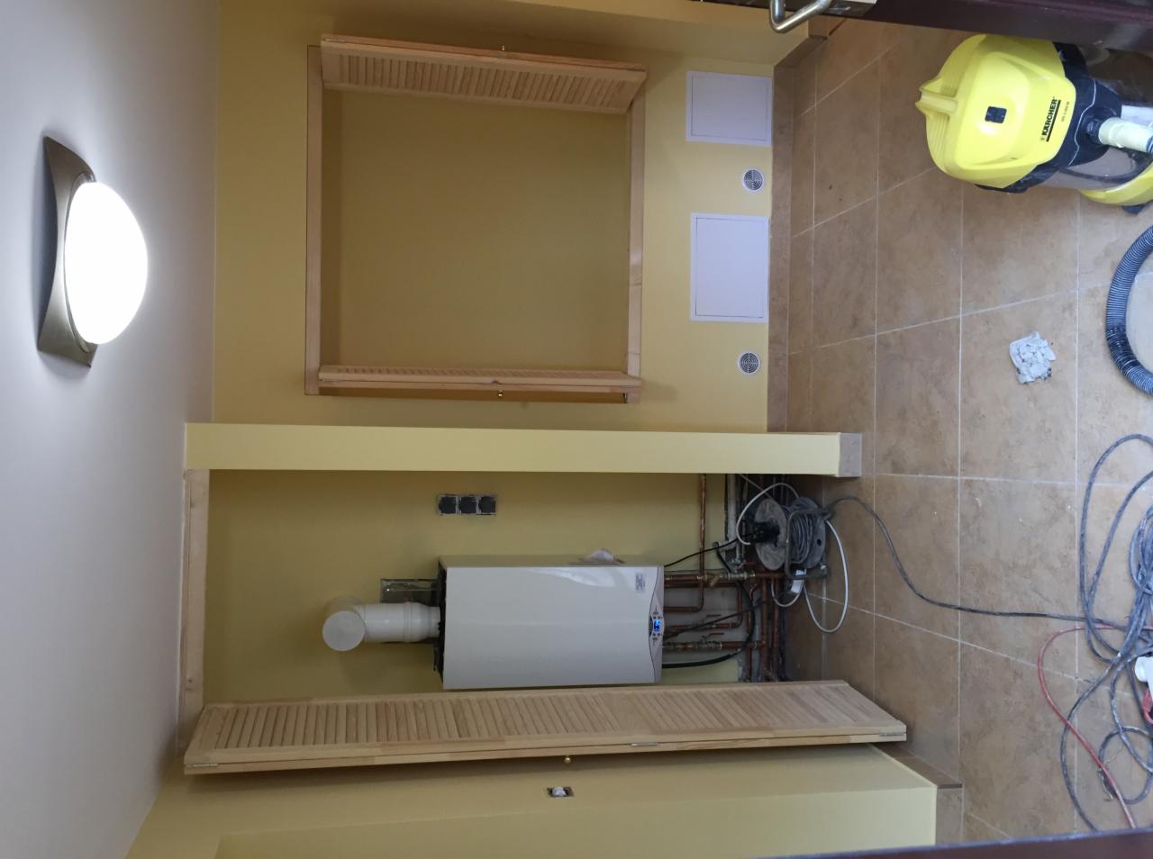 Żółta łazienka w trakcie montażu bojlera z widoczną instalacją hydrauliczną i elektryczną, z żółtym odkurzaczem Karcher w rogu i ramą szafki bez drzwi nad bojlerem.