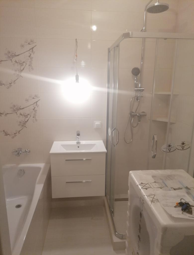 Wykończona łazienka w jasnych barwach: białe płytki z motywem kwiatowym, kabina prysznicowa ze srebrnymi okuciami, biała szafka z umywalką i baterią, wanna oraz pralka częściowo zakryta folią...