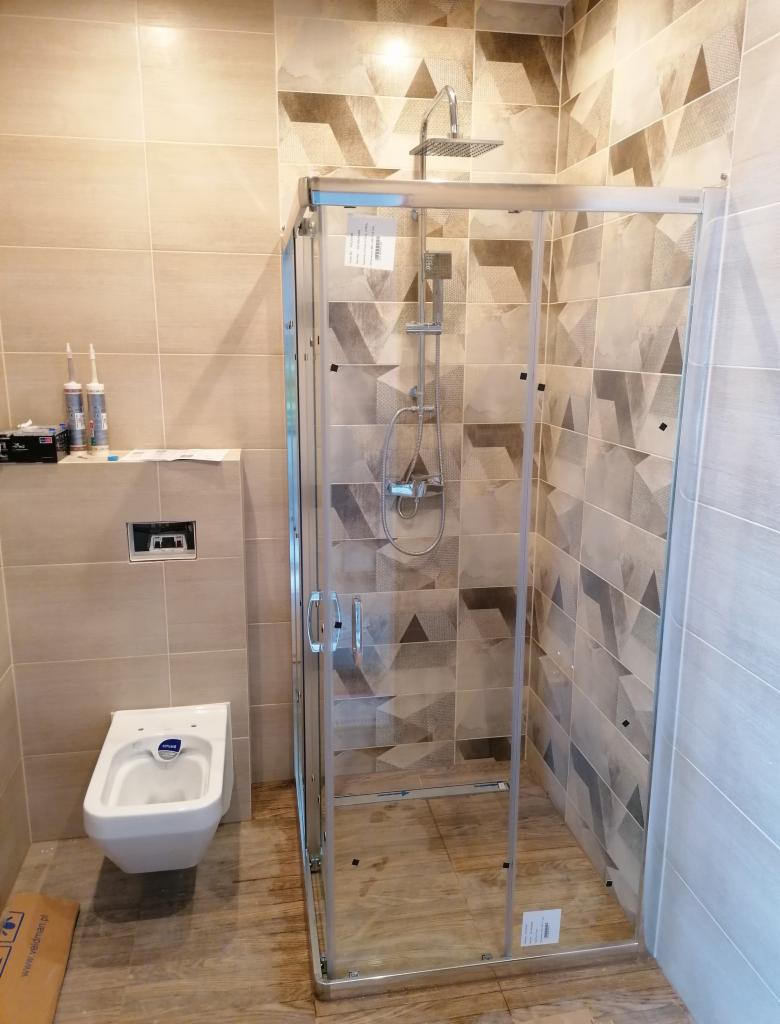 Nowoczesna łazienka po remoncie: kabina prysznicowa z deszczownicą, podwieszana toaleta i beżowo-brązowe płytki z geometrycznym wzorem.