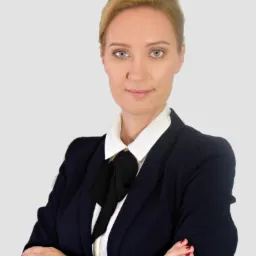 Portret kobiety w ciemnym garniturze i białej koszuli z czarną kokardą, z zegarkiem na ręce i skrzyżowanymi ramionami na jasnoszarym tle.