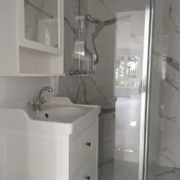 Biała umywalka z szafką i armaturą retro na tle kabiny prysznicowej z marmurowymi płytkami w łazience z oknem.