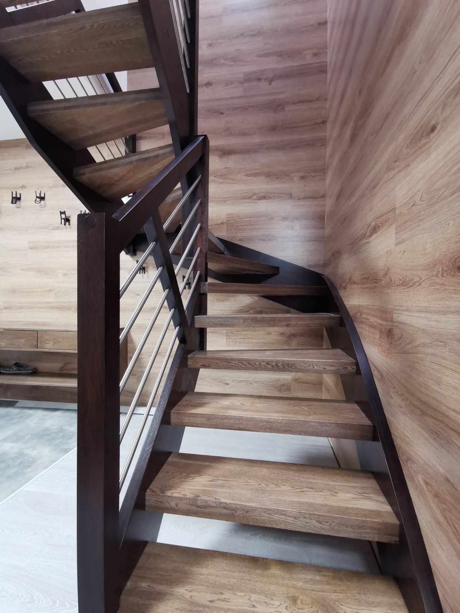 Drewniane schody z ciemnymi poręczami i metalowymi prętami, widok z dołu, ściana wykończona panelami imitującymi drewno.