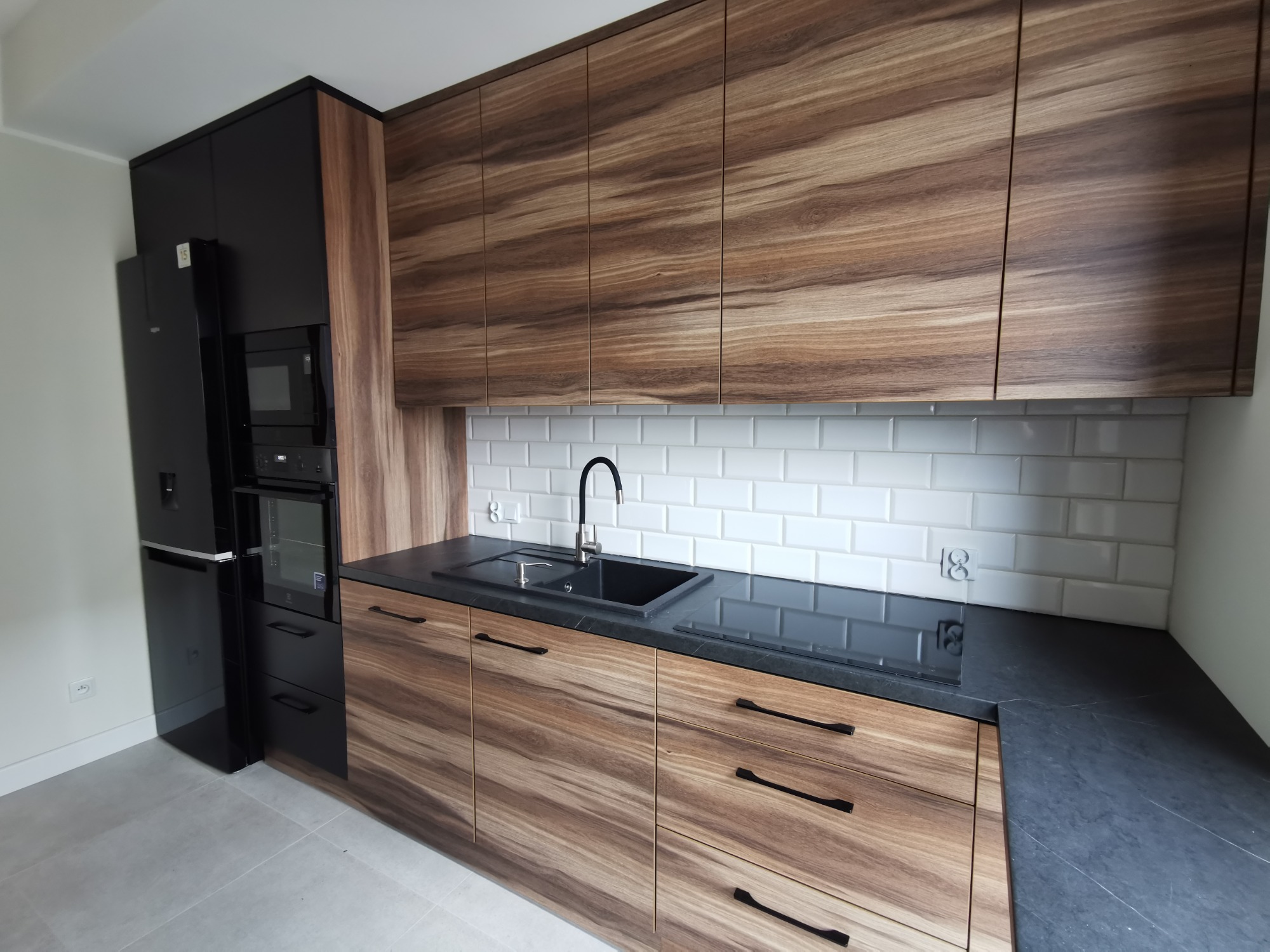 Nowoczesna kuchnia z drewnianymi frontami szafek w odcieniach brązu, czarnym blatem, białymi płytkami metra na ścianie, czarną armaturą i sprzętem AGD, minimalistyczne uchwyty.