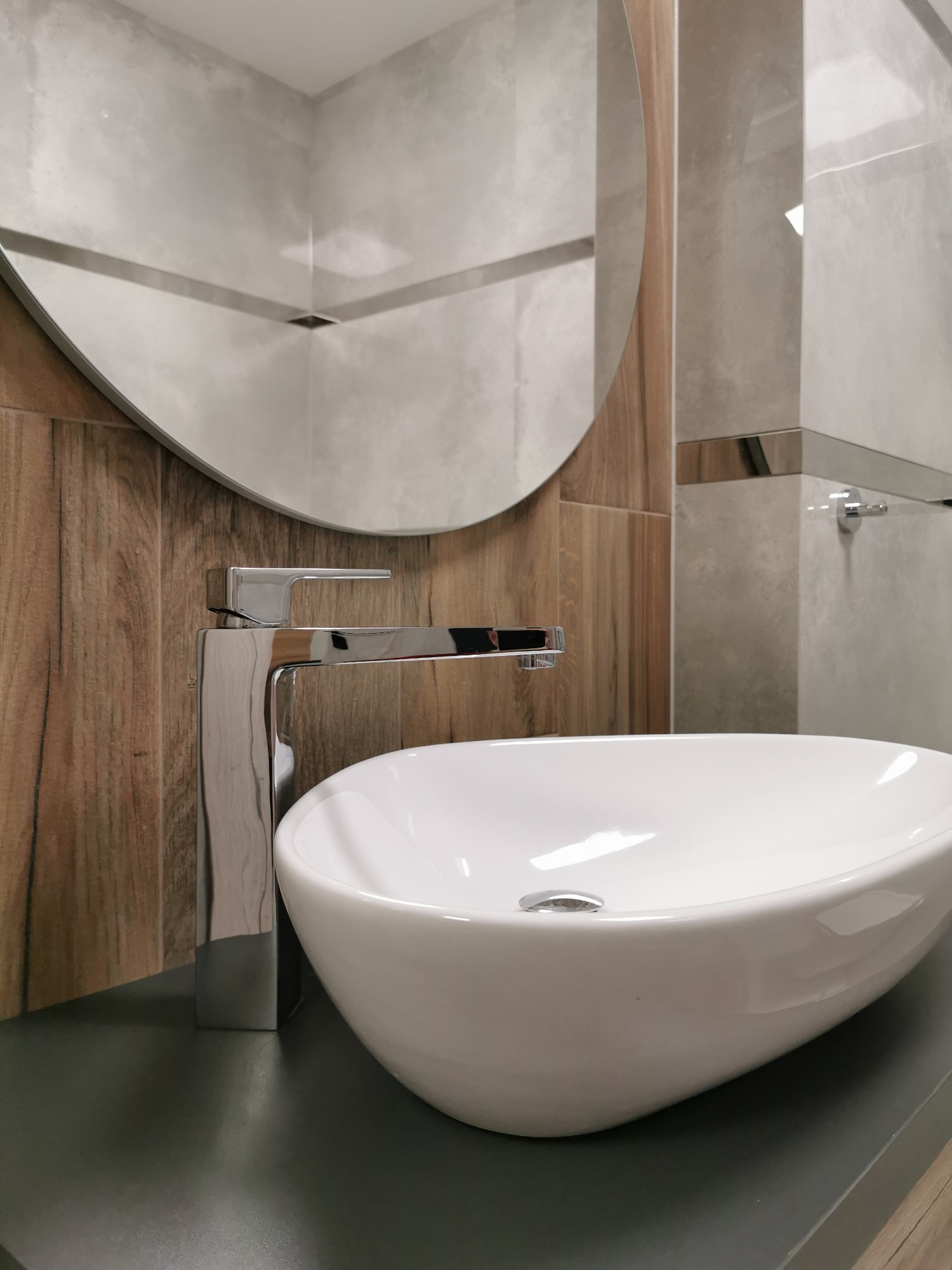 Biała, owalna umywalka nablatowa z chromowaną baterią na tle drewnopodobnych i szarych płytek, z okrągłym lustrem w tle, tworząca nowoczesną aranżację łazienki.