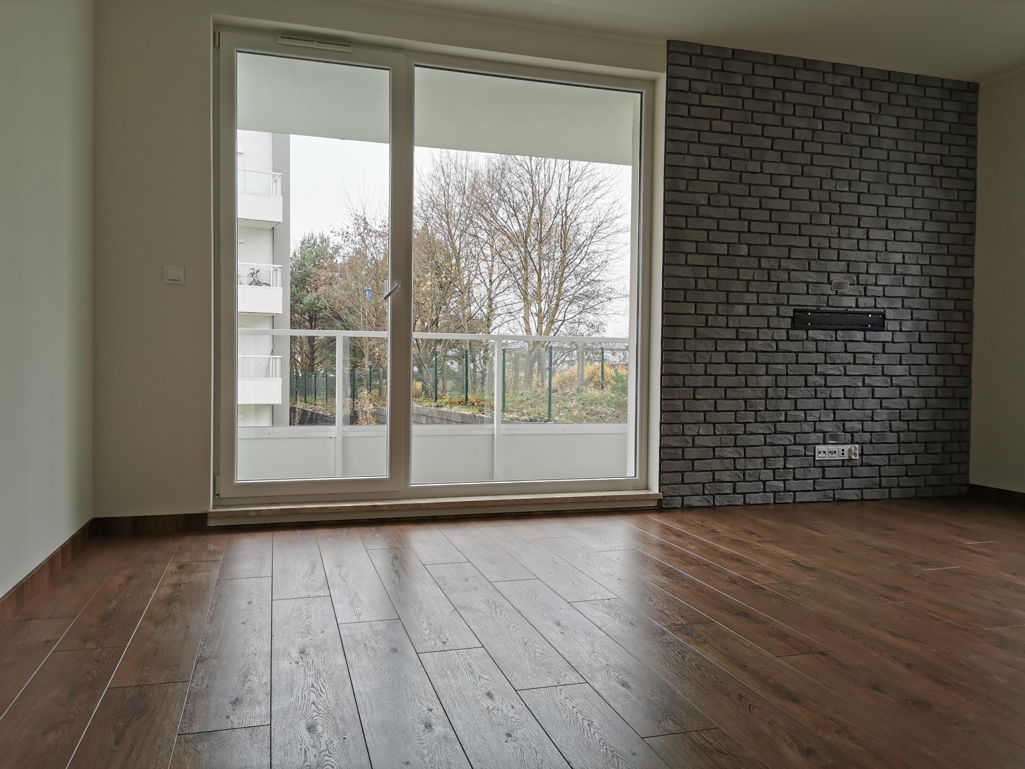 Pusty pokój z panelami podłogowymi w odcieniach brązu, ściana z szarej cegły dekoracyjnej z gniazdkami elektrycznymi i dużym oknem balkonowym z widokiem na drzewa.
