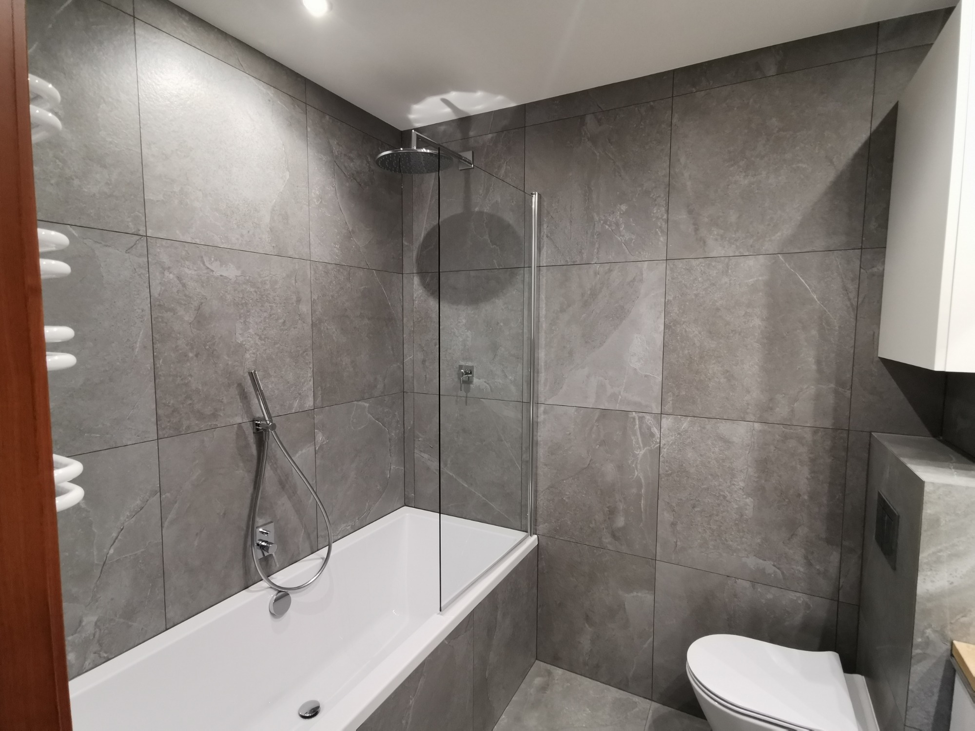 Nowoczesna łazienka z szarymi płytkami imitującymi kamień, białą wanną z parawanem, toaletą podwieszaną i grzejnikiem drabinkowym.