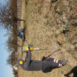 Mężczyzna w odzieży roboczej, z nausznikami ochronnymi, obsługuje wiertnicę glebową z żółtym silnikiem spalinowym na zewnątrz; w tle trawa, drzewa bez liści i częściowo widoczny niebieski samochód.