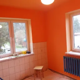 Pomarańczowe wnętrze pokoju po świeżym malowaniu, z zabezpieczoną folią podłogą, grzejnikiem i oknami, widoczny stołek i narzędzia malarskie na parapecie.