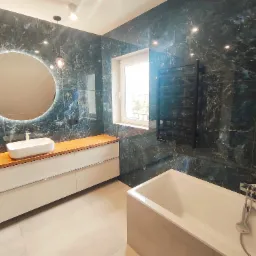 Wykończona łazienka z ciemnozielonymi płytkami imitującymi marmur, podświetlanym okrągłym lustrem, białą umywalką nablatową na drewnianym blacie, grzejnikiem drabinkowym i białą wanną.