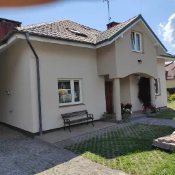 Jasny, dwukondygnacyjny dom z nową elewacją, brązowym dachem i drewnianą ławką przed oknem, sfotografowany w słoneczny dzień.