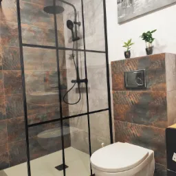 Nowoczesna łazienka z kabiną prysznicową typu walk-in z czarną armaturą, płytkami imitującymi rdzę, wiszącą toaletą i dekoracjami roślinnymi na szafce.
