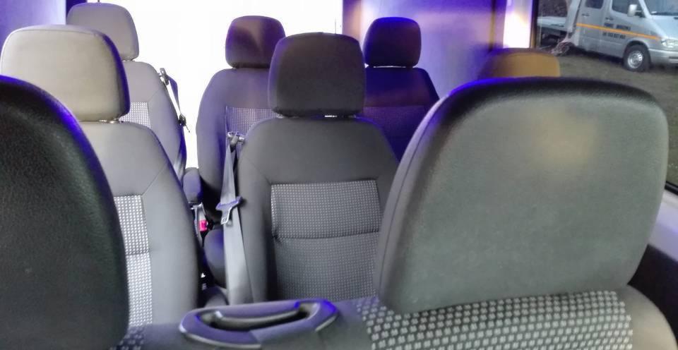 Wnętrze busa z rzędami szarych i czarnych foteli z materiałową tapicerką, widoczny srebrny bus na zewnątrz.