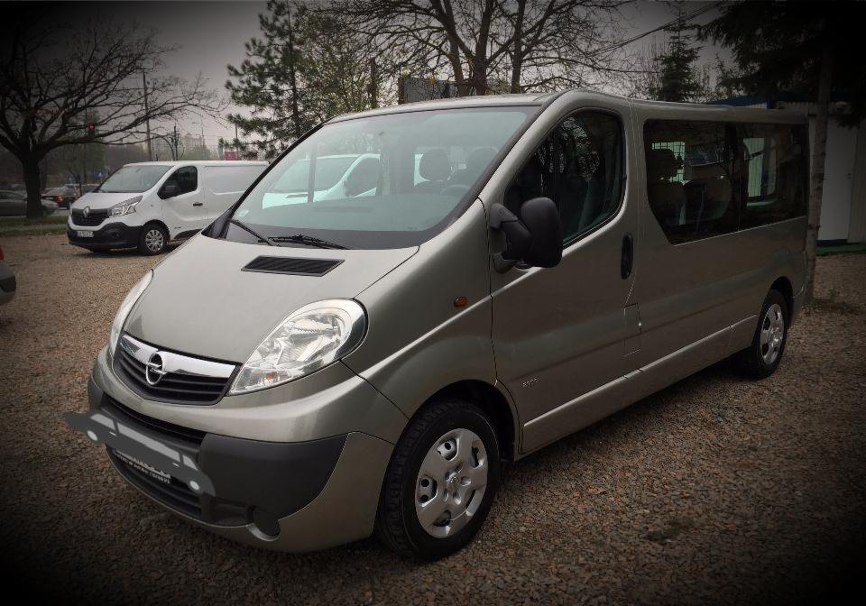 Szary bus Opel Vivaro na żwirowym parkingu, z widocznymi innymi pojazdami w tle.