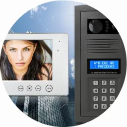 Zestaw wideodomofonowy: panel zewnętrzny z kamerą i klawiaturą numeryczną Elfon oraz monitor wewnętrzny z wyświetlaczem, tło rozmazane z motywem architektury miejskiej.