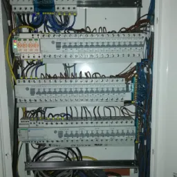 Otwarta skrzynka elektryczna z licznymi bezpiecznikami, przewodami w różnych kolorach i listwami zaciskowymi, widoczne elementy marki EATON i ETI, z zieloną diodą wskazującą stan pracy.