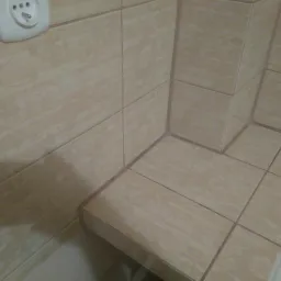 Beżowe płytki ceramiczne z delikatnym wzorem kwiatowym, ułożone w narożniku łazienki, widoczny fragment gniazdka elektrycznego i krawędź wanny.