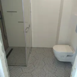 Nowoczesna łazienka z kabiną prysznicową typu walk-in, wiszącą toaletą i podłogą w czarno-białe wzory geometryczne, widok z perspektywy wejścia, częściowo wykończona.