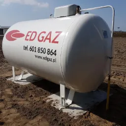 Biały, poziomy zbiornik gazowy z logo ED GAZ i danymi kontaktowymi, umieszczony na betonowych fundamentach na placu budowy.
