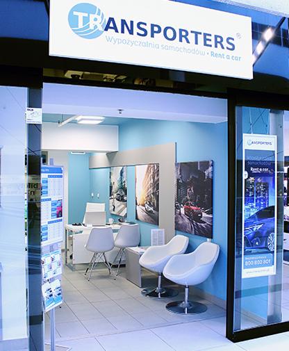 Jasne wnętrze biura z białymi krzesłami i plakatami samochodów, widoczne przez szklane drzwi z napisem 'Transporters - Wypożyczalnia samochodów - Rent a car' nad wejściem.
