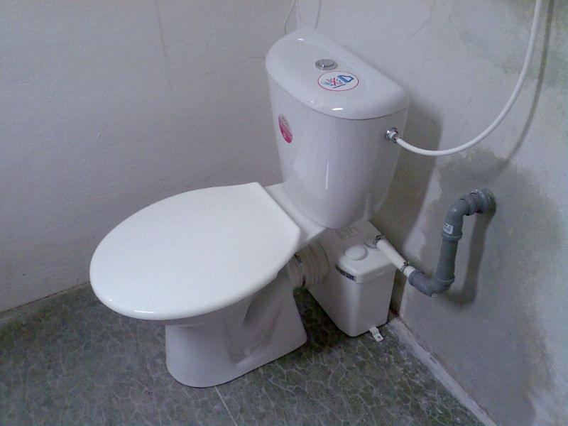 Biała toaleta z zamontowanym maceratorem i szarą rurą odpływową w rogu łazienki z szarymi płytkami podłogowymi.