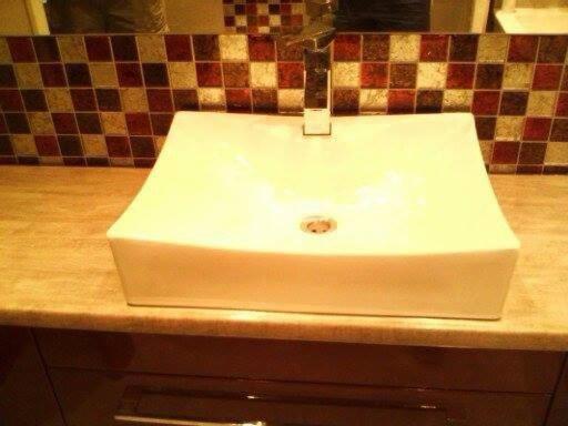 Prostokątna, biała umywalka nablatowa z chromowaną baterią, zamontowana na blacie z imitacji drewna, tło stanowi mozaika z płytek w odcieniach brązu i beżu.