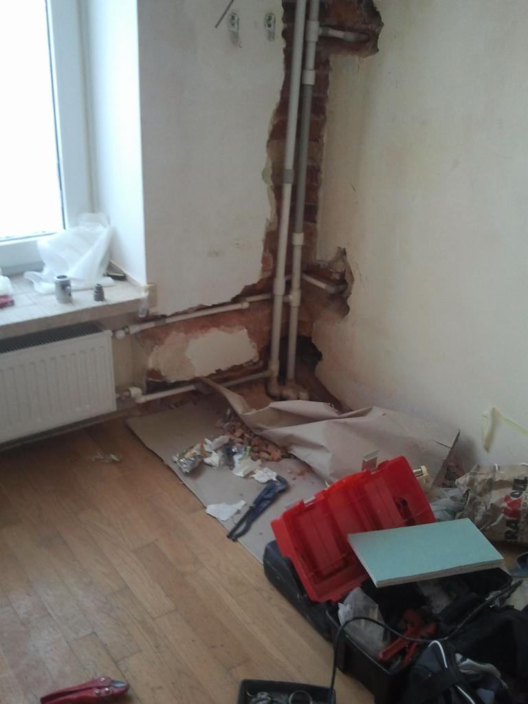 Odkryte rury wodne w ścianie podczas remontu, widoczne narzędzia i materiały budowlane na podłodze, częściowo wykończone ściany i okno w tle.