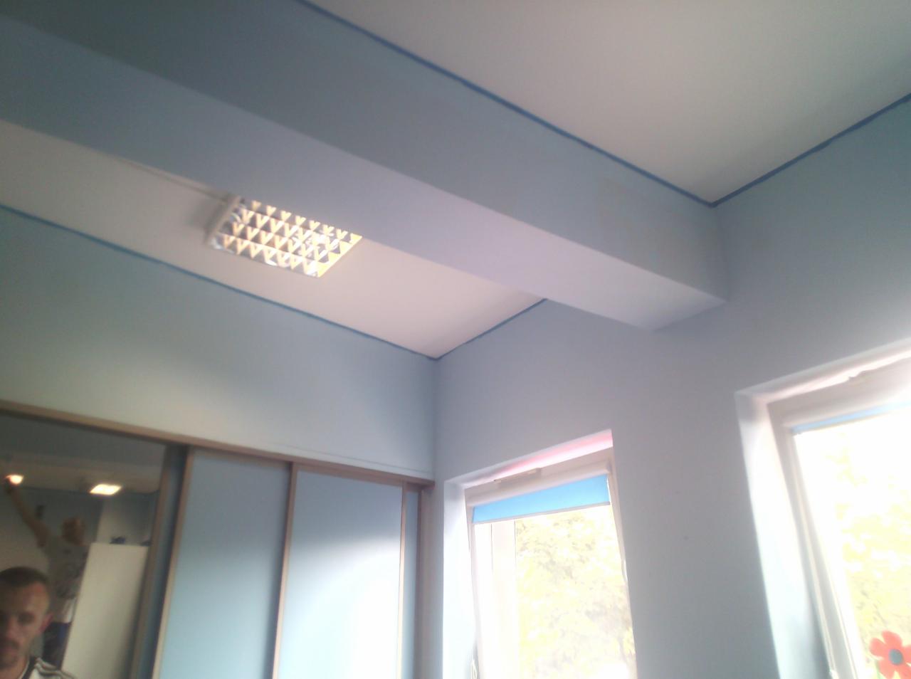 Pomieszczenie po malowaniu ścian na jasny błękit, widoczne świeżo pomalowane ściany, sufit z lampą rastrową, okno z roletą i fragment szafy przesuwnej z lustrem.