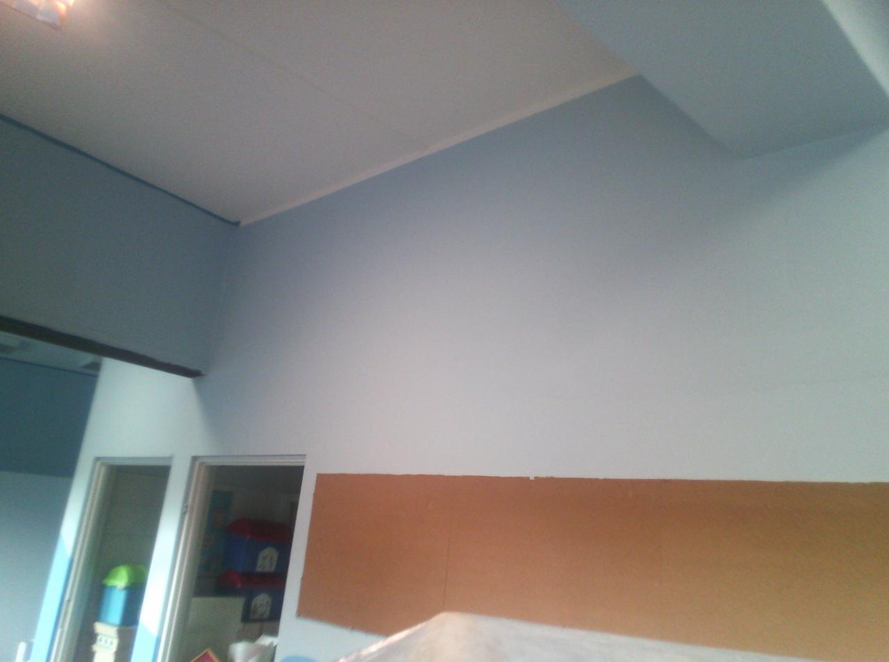 Świeżo pomalowana ściana na jasny odcień niebieskiego, z widocznym brązowym fragmentem ściany przygotowanym do dalszych prac, widoczne drzwi do innego pomieszczenia